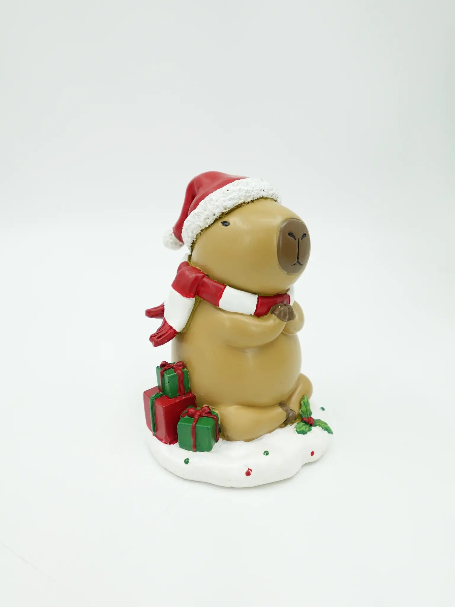A holiday capybara figurine wearing a Santa hat and matching scarf, sitting on a snowy base with gift boxes around it