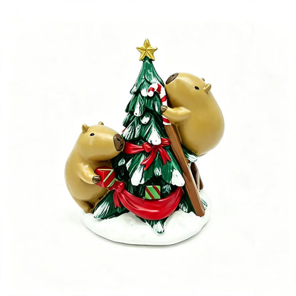 A festive resin figurine of two tan capybaras decorating a green Christmas tree with a red bow and gifts, set on a white snowy base against a white background.