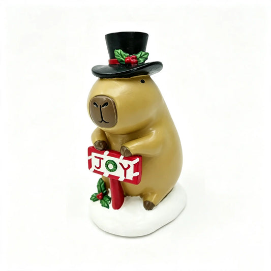 Hand-painted Christmas capybara figurine in black top hat with holly, holding a red JOY sign on a snowy white base, front view on white background